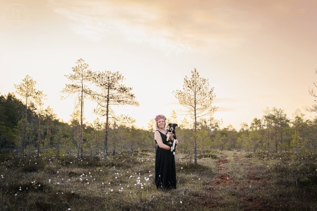 mustaan mekkoon ja kukkaseppeleeseen pukeutunut vaaleahiuksinen nainen pitelee sylissään mustavalkoista bordercolliepentua suolla aamulla auringonnousun aikaan, runsaasti tupasvilloja taustalla